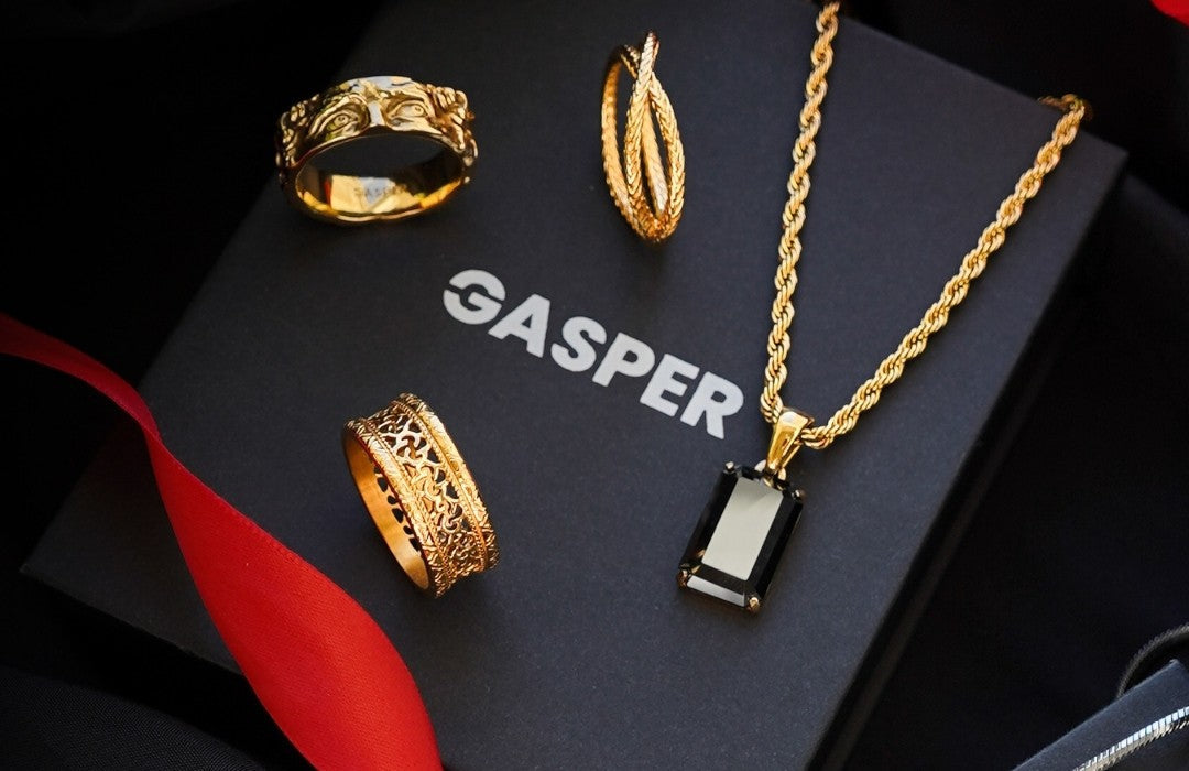 Gold rings and a black onyx pendant on a gold rope chain laid on a Gasper gift box with a red ribbon
